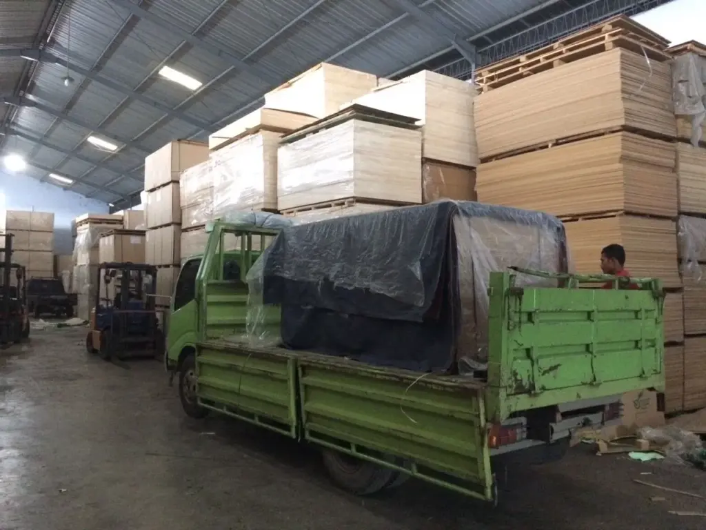 Aktivitas pengiriman truk di gudang stok triplek Indoho.com yang penuh dengan tumpukan plywood dan bahan kayu olahan siap kirim. Menunjukkan kapasitas distributor besar untuk kebutuhan proyek di Jakarta dan seluruh Indonesia.