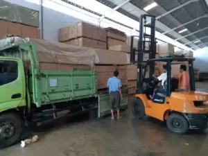 Forklift sedang memuat tumpukan kotak plywood ke atas truk kuning di gudang distributor PT Indoho Santosa Abadi Semarang.